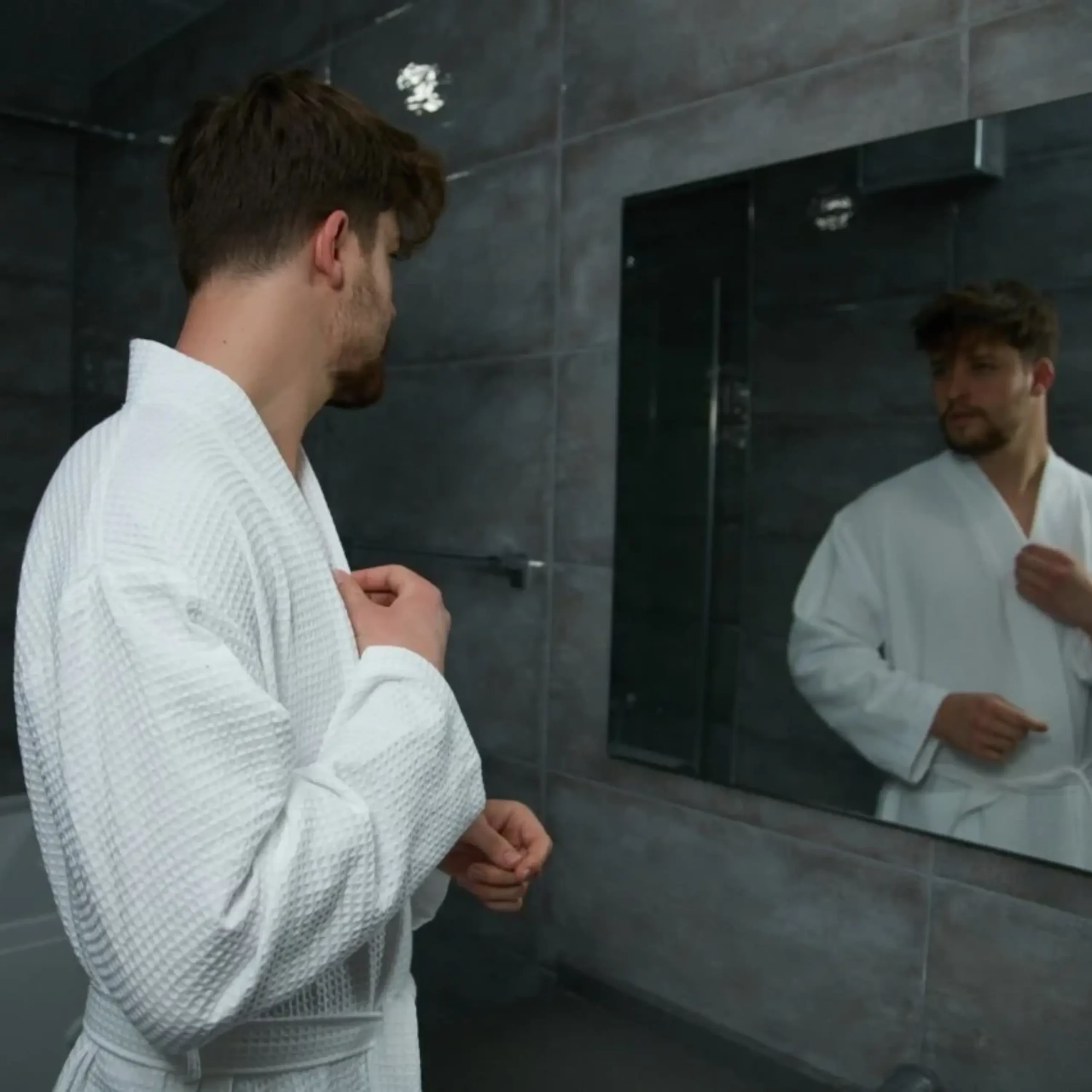 Man in a white waffle pique robe looking in a bathroom mirror