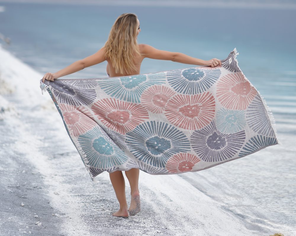 Woman walking along a turquoise shore holding an open Bagno Milano peshtemal beach towel